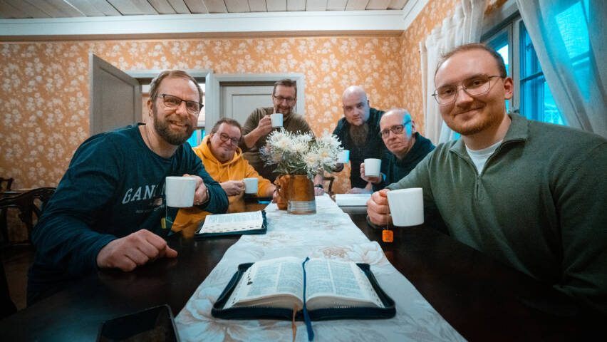Miehet nostavat kahvikuppeja pöydän ympärillä ja hymyilevät.